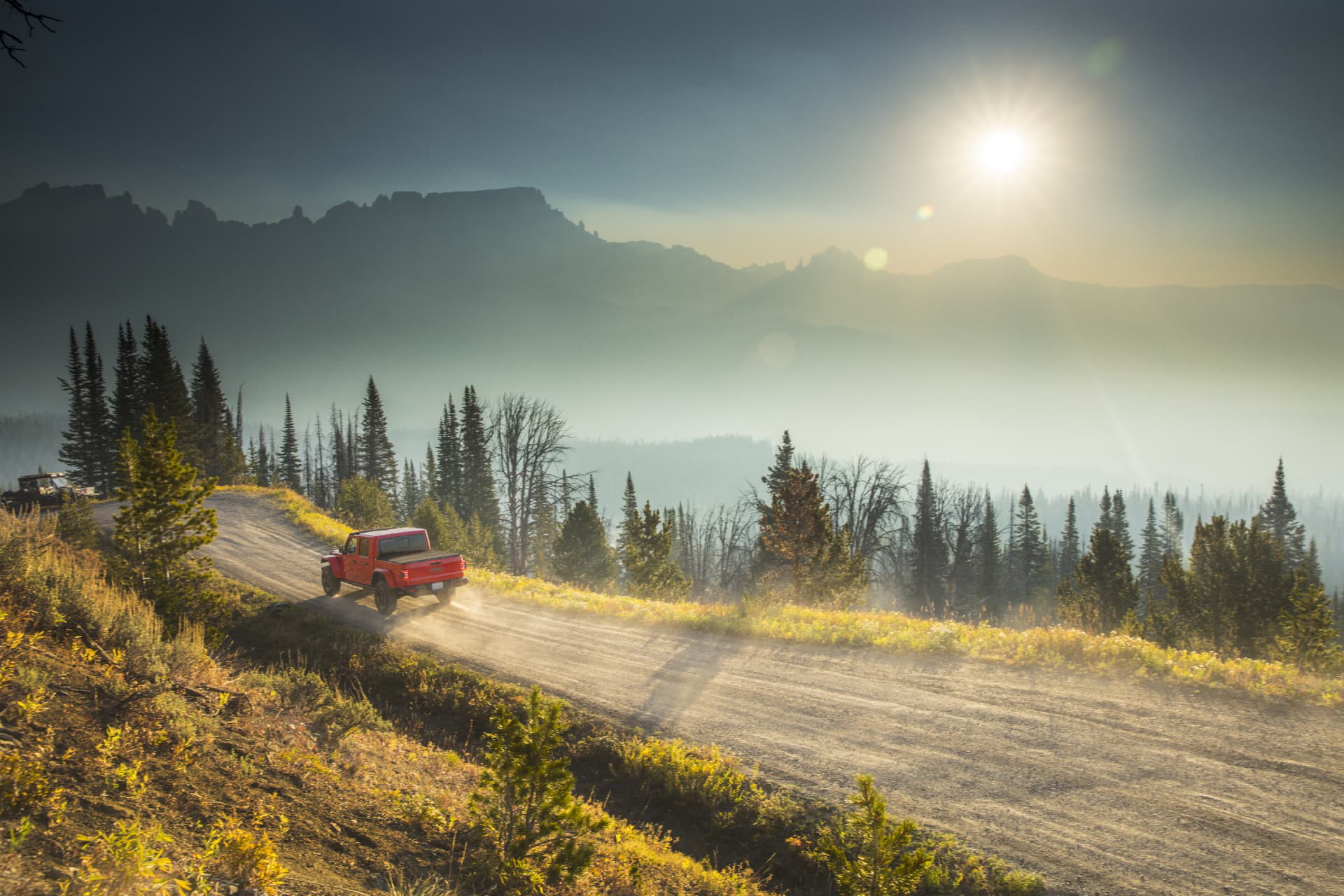 Un Jeep Gladiator rojo circulando por un paisaje montañoso al atardecer.