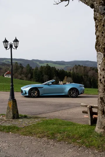 Vista lateral del Aston Martin DB12 Volante 2024 aparcado en entorno rural.