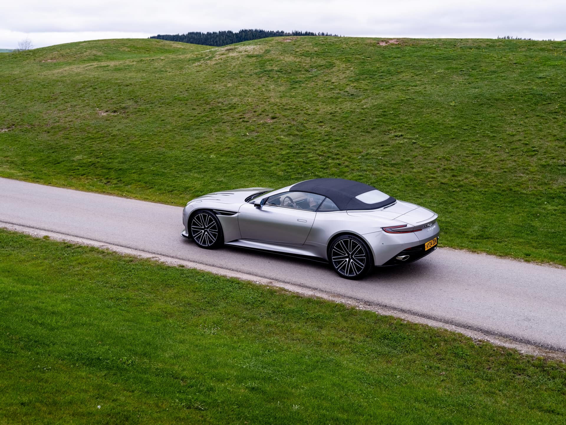 Vista lateral del Aston Martin DB12 Volante 2024 deslizándose elegancia sobre carretera campestre.