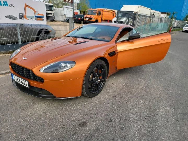 Un Aston Martin en tono naranja con la puerta abierta, mostrando su perfil imponente.