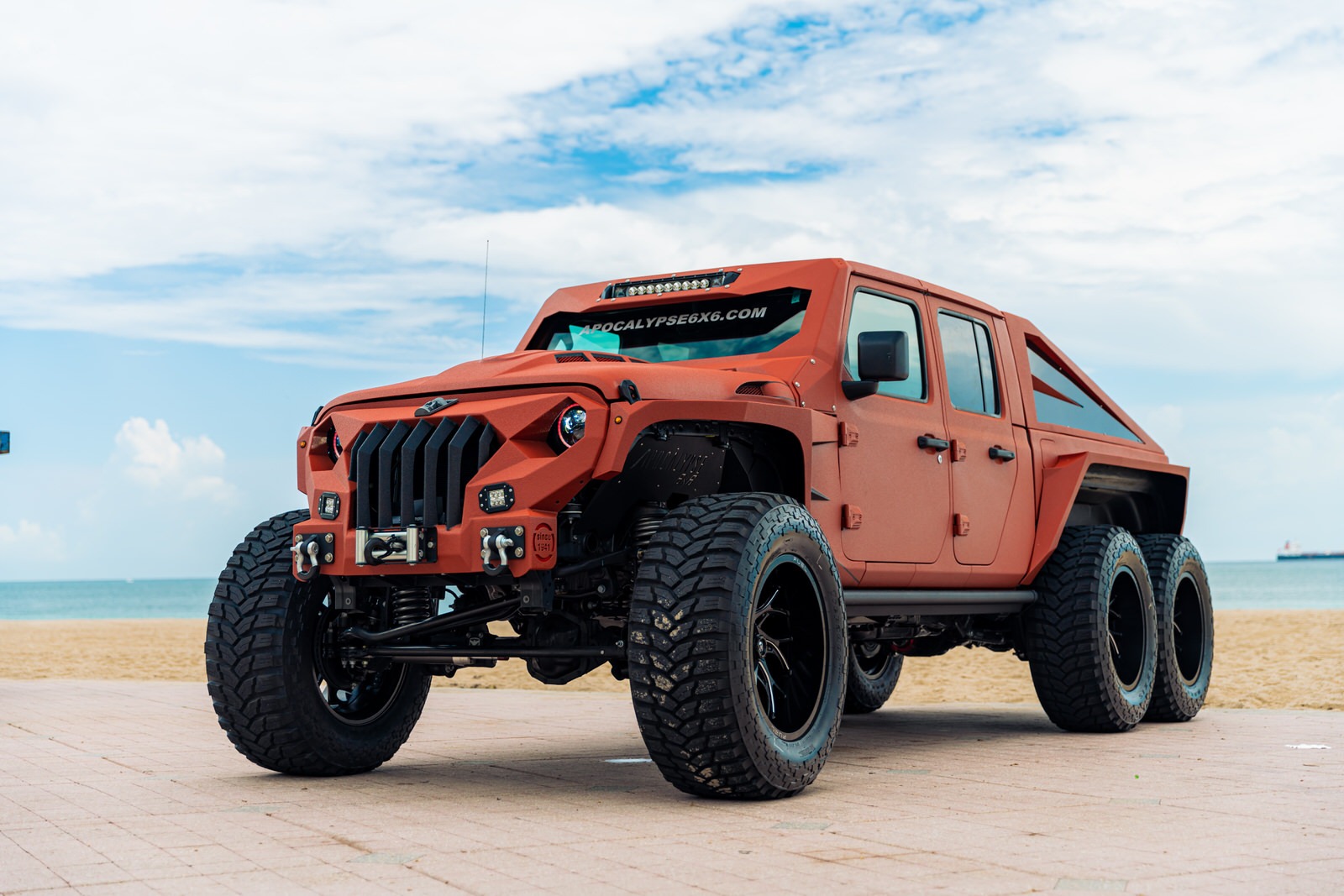 Un poderoso Jeep 6x6 posa majestuoso en la playa, pintura naranja y llantas imponentes.
