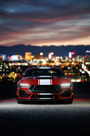 Mustang rojo con franjas blancas y negras, luces encendidas, fondo urbano nocturno.