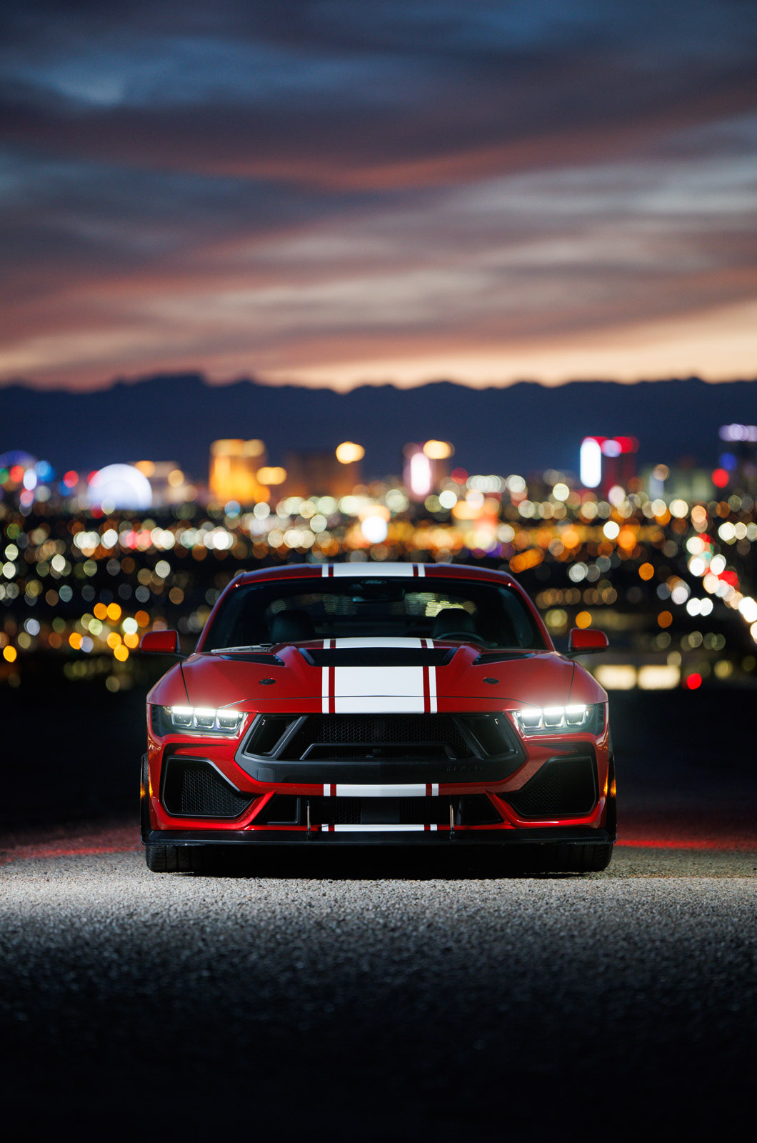 Mustang rojo con franjas blancas y negras, luces encendidas, fondo urbano nocturno.