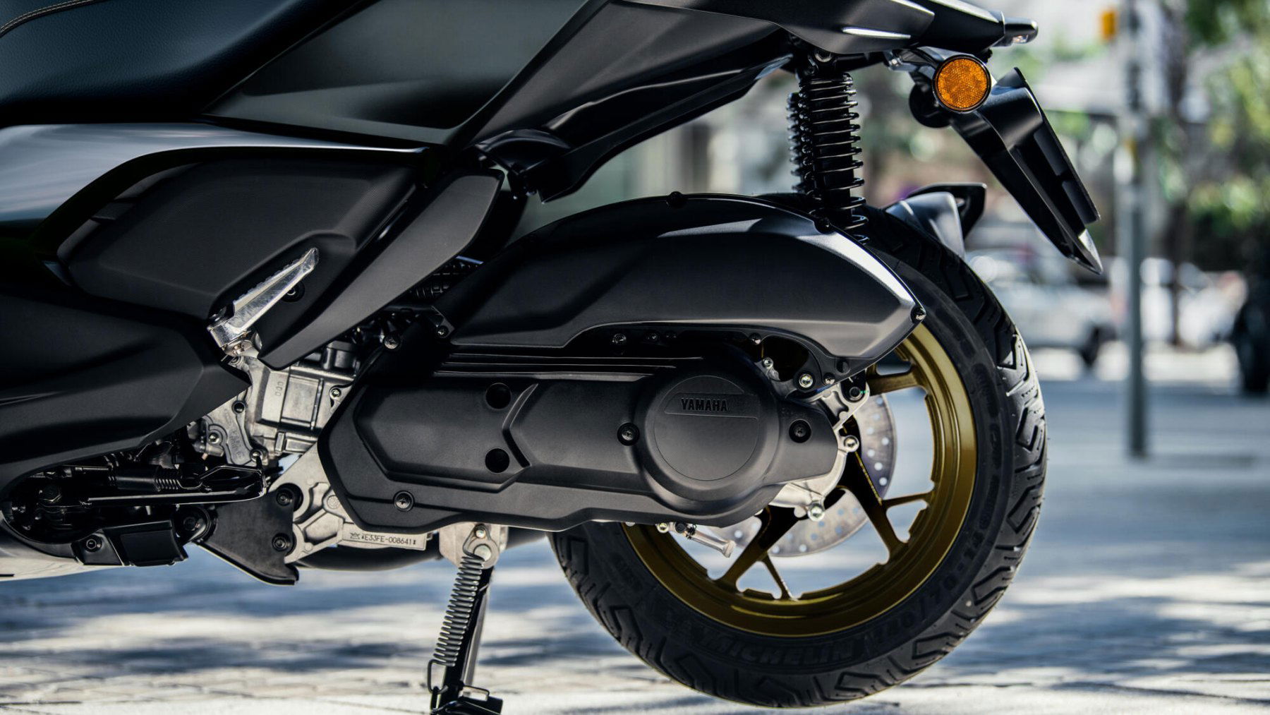 Vista detallada del sistema de suspensión y la rueda trasera dorada de la Yamaha X-Max 125cc. Vista detallada del sistema de suspensión y la rueda trasera dorada de la Yamaha X-Max 125cc.