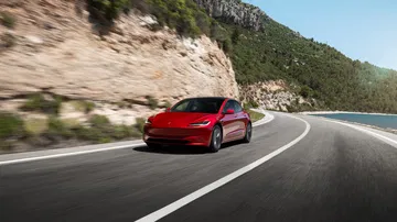 Vista lateral de un Tesla Model 3 rojo circulando por carretera montañosa.