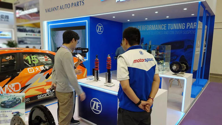 Exposici&oacute;n con empleados junto a stand de suspensiones ZF, coche naranja al fondo.