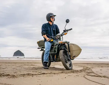 Vista lateral de la UBCO 2X2 eBike en ambiente playero con tabla de surf adjunta