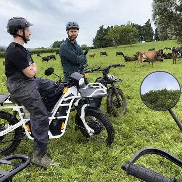 Dos individuos equipados con cascos sobre una motocicleta eléctrica UBCO 2x2 en terreno verde.