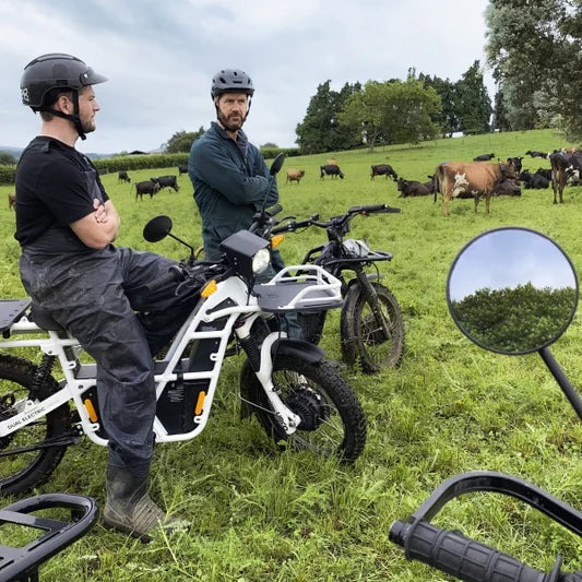 Dos individuos equipados con cascos sobre una motocicleta eléctrica UBCO 2x2 en terreno verde.