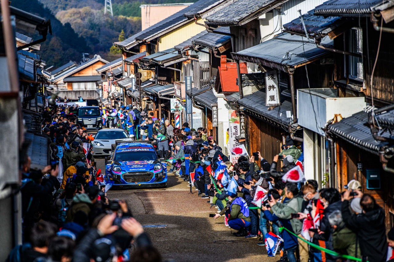 Un coche de rally compitiendo en el entorno histórico de Japón, rodeado de espectadores.