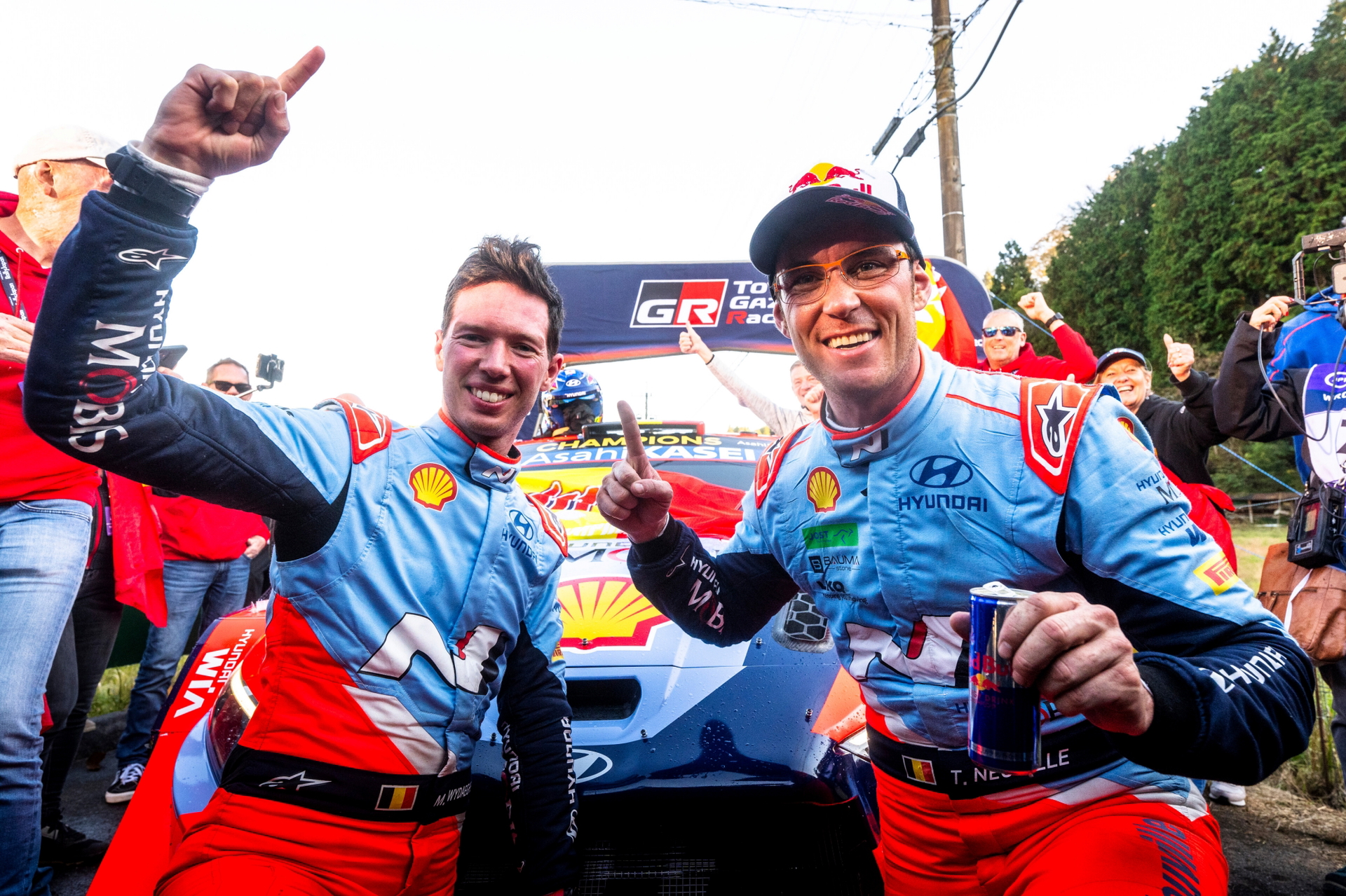 Dos pilotos celebran su victoria con un coche de carreras al fondo.